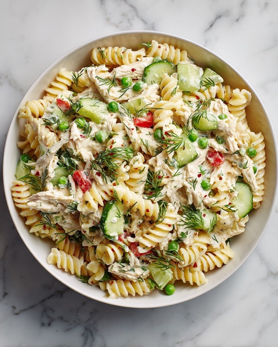 A close-up view of a pasta salad in a deep white bowl sitting on a white marbled surface, showing three main layers: pale yellow spiral rotini pasta forming the base with a soft texture, mixed with bright green peas scattered throughout as the second layer, and fresh cucumber cubes with light green skin and red bell pepper chunks adding pops of color on top. The whole salad is coated in a creamy white dressing dotted with small green herb pieces, likely dill, sprinkled evenly over the dish giving a fresh look. photo taken with an iphone --ar 4:5 --v 7 — Creamy Healthy Tuna Pasta Salad, healthy tuna pasta salad, nutritious seafood pasta dish, quick tuna pasta salad, wholesome summer pasta salad